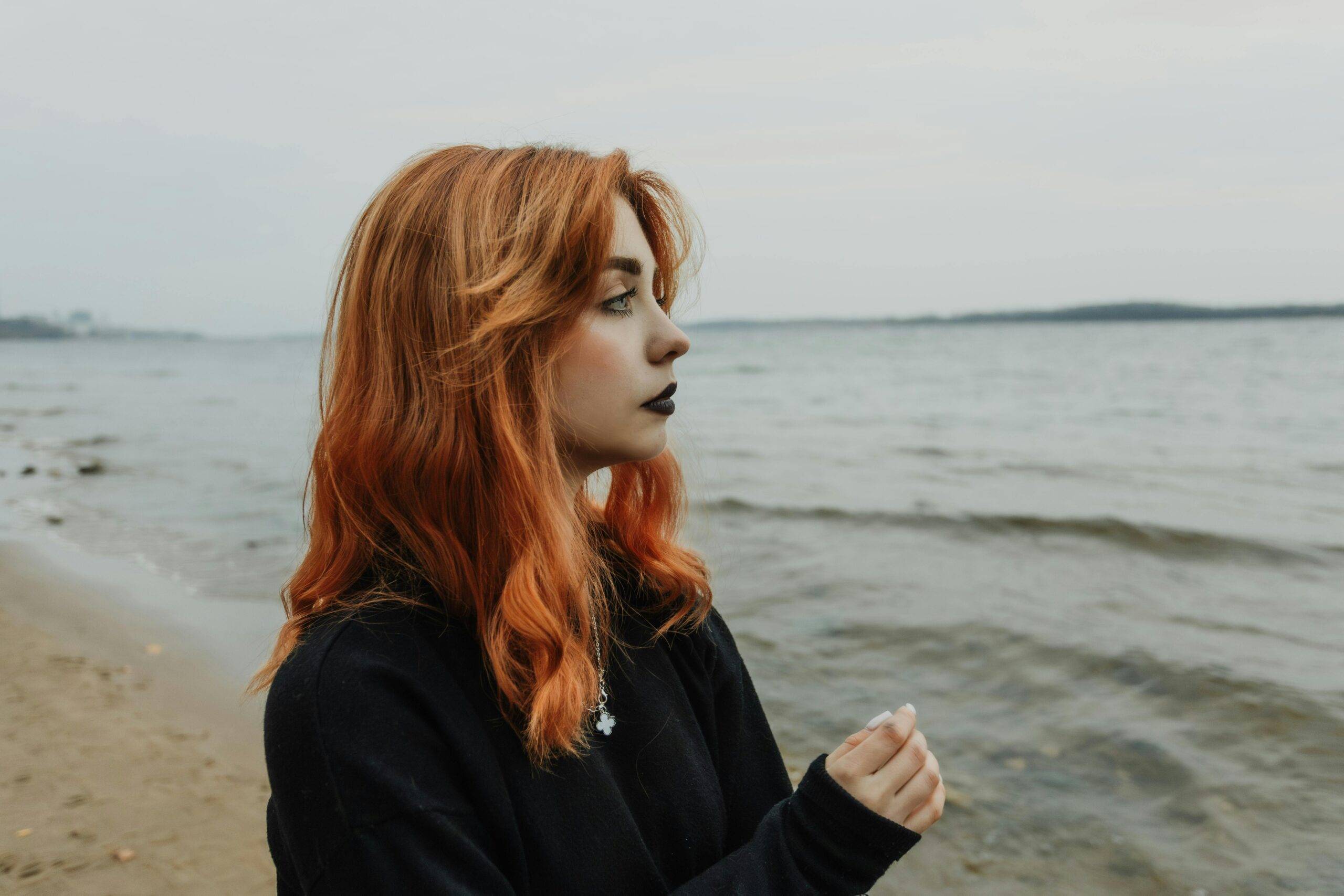 A contemplative young woman with red hair stands by the beach on an overcast day.